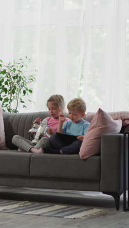 preschooler girl holds airplane toy while toddler boy looks in tablet. siblings sit on sofa near big window watching educational lessons on gadget