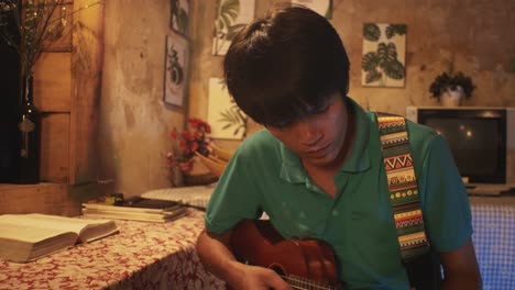 young asian male playing a ukulele while sitting on a chair in a rustic room