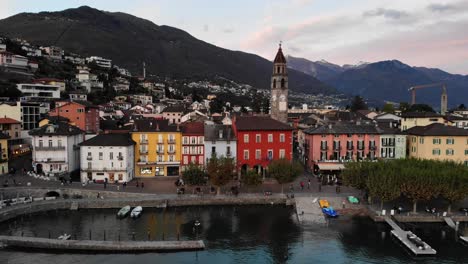 Sobrevuelo-Aéreo-A-Lo-Largo-Del-Paseo-Junto-Al-Lago-De-Ascona,-Ticino-A-Orillas-Del-Lago-Maggiore-En-La-Suiza-Italiana-Al-Final-De-Un-Día-De-Verano-Con-Casas-Coloridas-Y-La-Torre-De-La-Iglesia-A-La-Vista