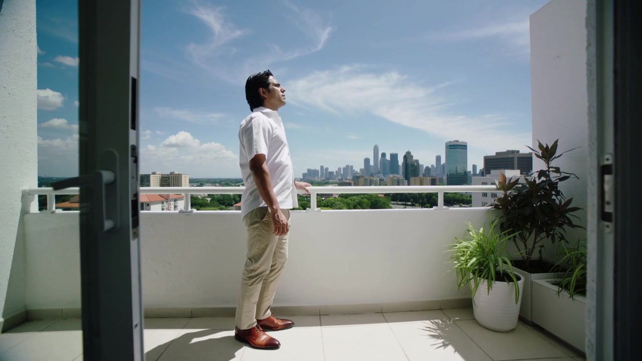 Uomo che cammina, camicia bianca, pantaloni beige, entra in balcone, raggiunge la ringhiera, osserva lo skyline con occhiali da sole