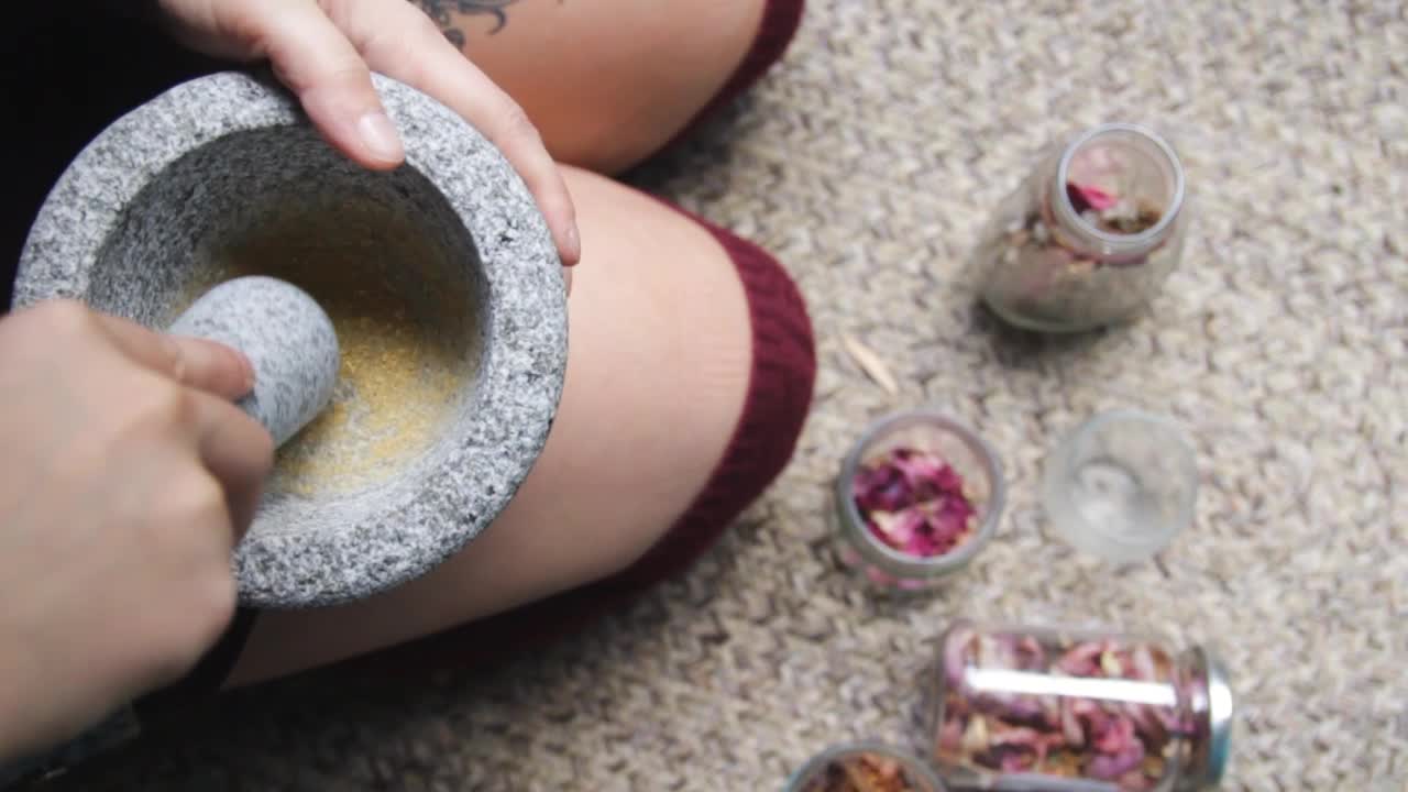 Straight down view of a female adult sitting on the ground, holding mortar bowl and pestle, crushing hard on flower petals to grind and extract organic yellow pigments to make water-colour paints.