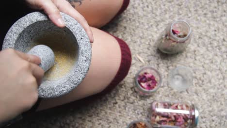straight down view of a female adult sitting on the ground, holding mortar bowl and pestle, crushing hard on flower petals to grind and extract organic yellow pigments to make water-colour paints.