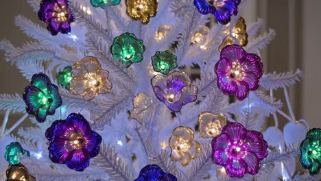 montage showcasing a white christmas tree decorated with iridescent flower ornaments in vibrant purple, green, and gold hues, illuminated by small lights, creating a festive and elegant atmosphere