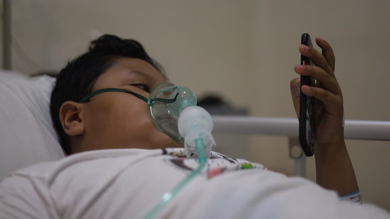 niño asiático de 9 años con máscara de oxígeno en la cara en la cama del hospital mientras juega al teléfono celular