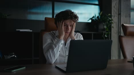 a tired guy with curly hair in a white shirt looks tiredly at a gray laptop and holds his head with his hands during a hard day at work in the office