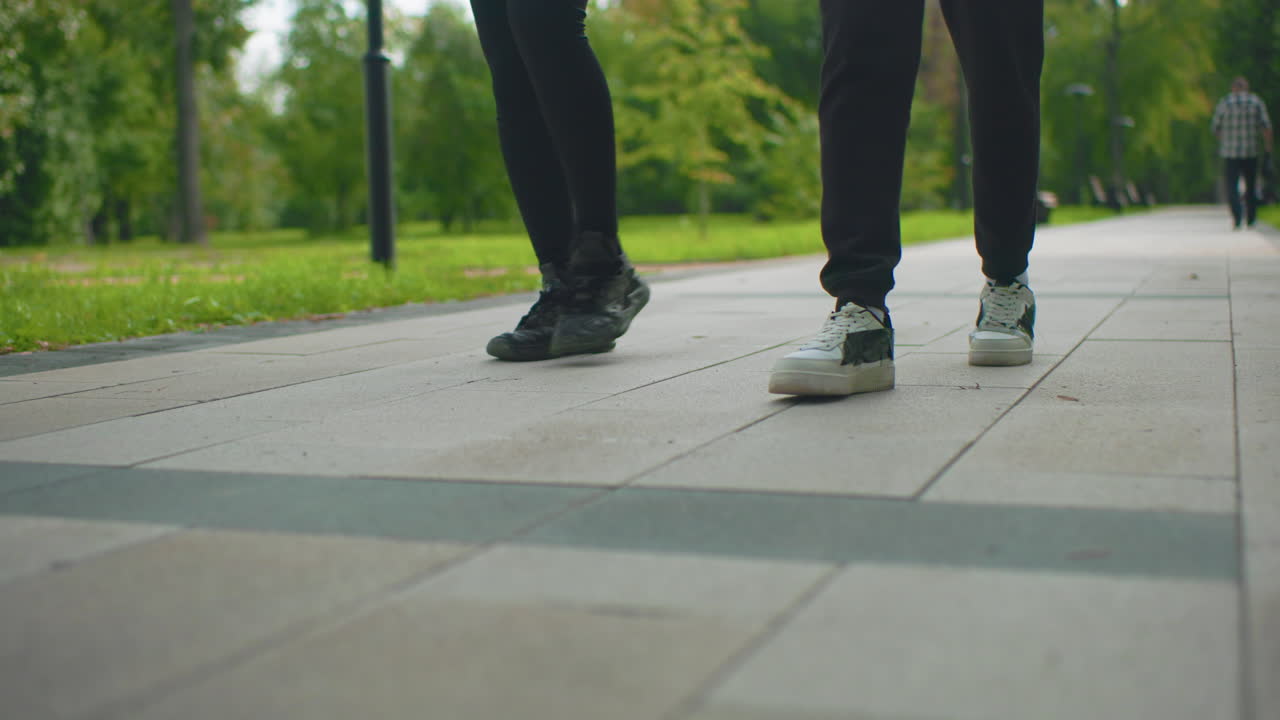 closeup feet walking on tiled park path, paired slow stroll past benches and leafy greens, soft evening light and gentle bokeh, contemplative pace with casual sneakers, intimate lowangle framing