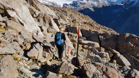 female hiker navigates boulder field with snow covered mountains in background