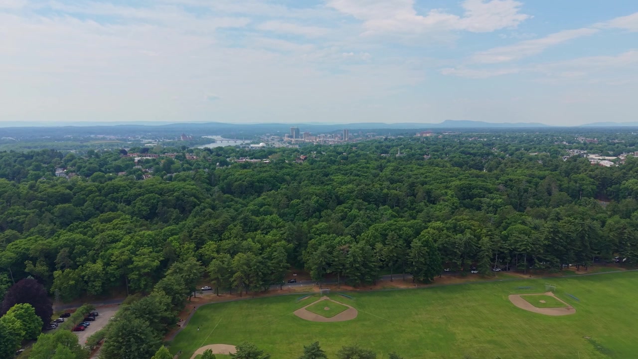 Vista lejana de la ciudad de Springfield y el río Connecticut desde un parque arbolado con campo de béisbol en Massachusetts. Toma amplia con dron.