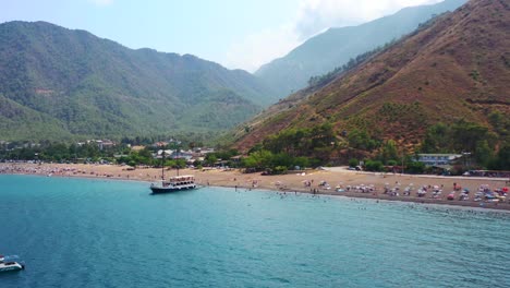 Luftdrohnenaufnahme-Des-Strandes-Von-Adrasan-Voller-Menschen,-Während-Ein-Boot-Am-Strand-Vor-Anker-Liegt-Und-An-Einem-Heißen-Sommertag-In-Der-Trockenen-Berglandschaft-Der-Türkei-Auf-Touristen-Wartet