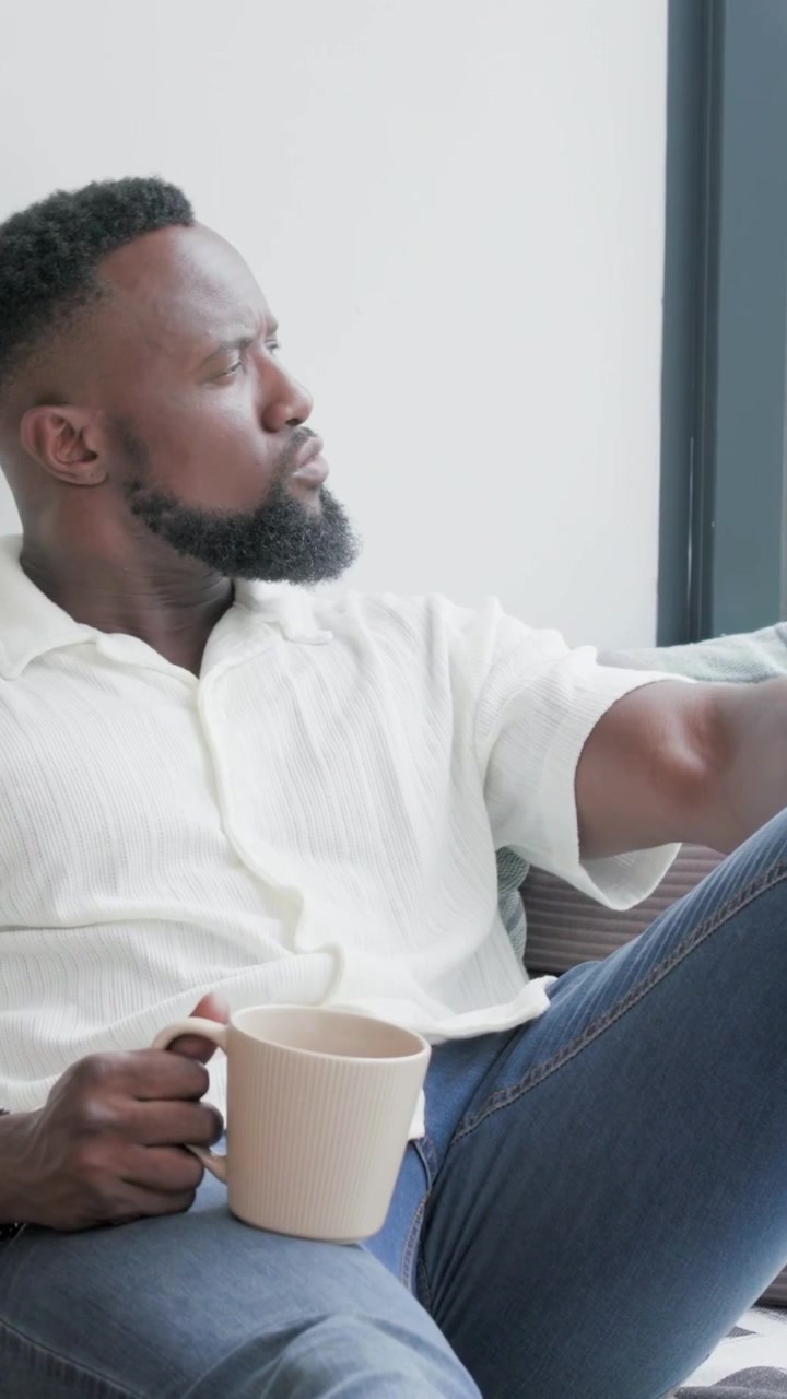 Vertical video: Sipping man in light shirt and blue jeans on couch by window, holding beige mug