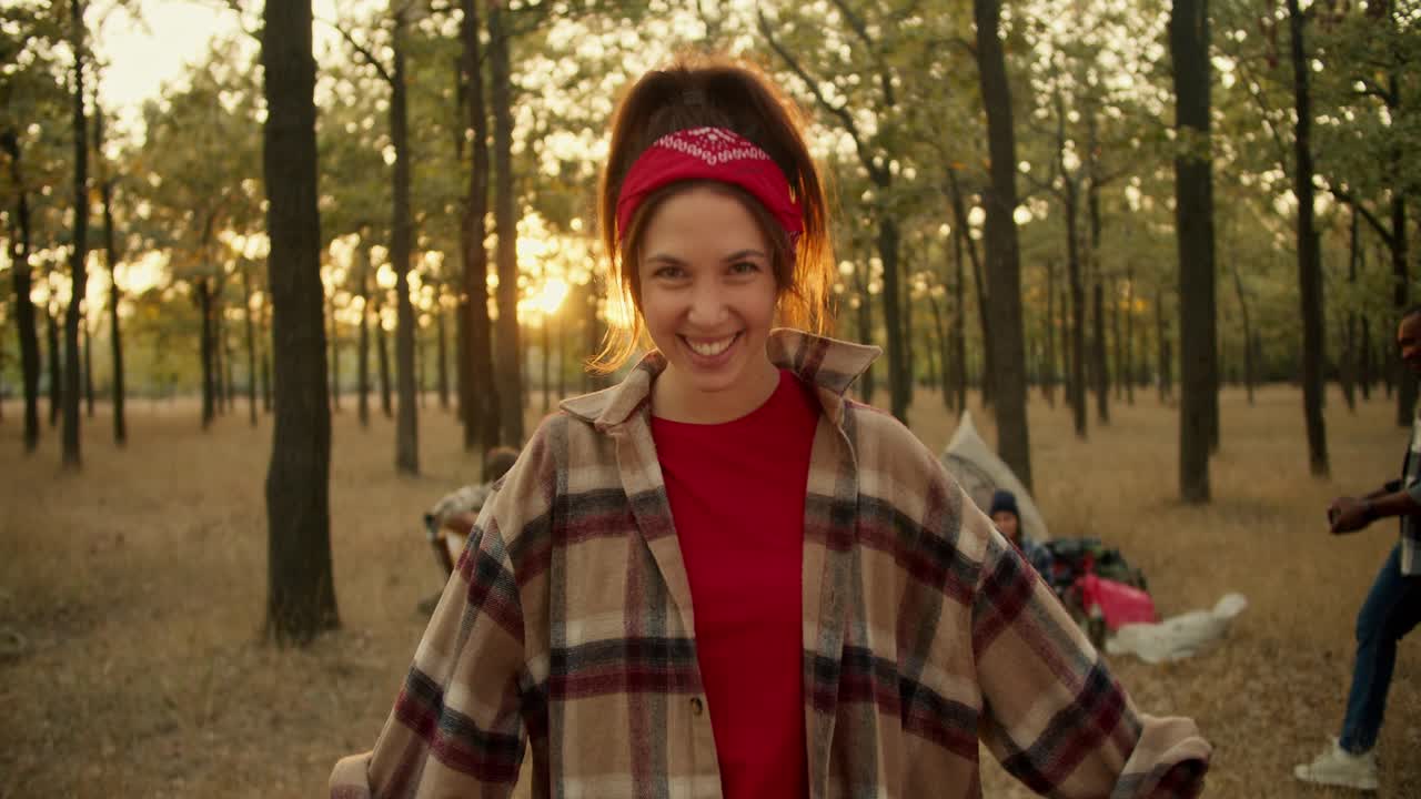 retrato de una chica feliz con una camisa a cuadros que se endereza el cabello y la bandana y se para, sonriendo y regocijándose contra el fondo del resto del grupo de excursionistas y tiendas de campaña en el verde bosque soleado de verano