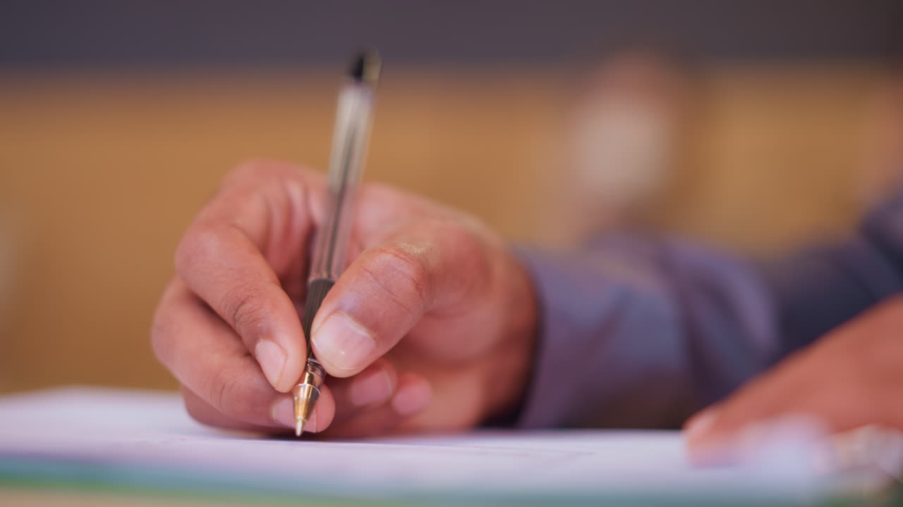 Close-up hand gripping pen while writing on paper, detailed focus on motion and pen tip, individual engaged in office task or academic work, warm blurred background suggests calm, productive atmosphere