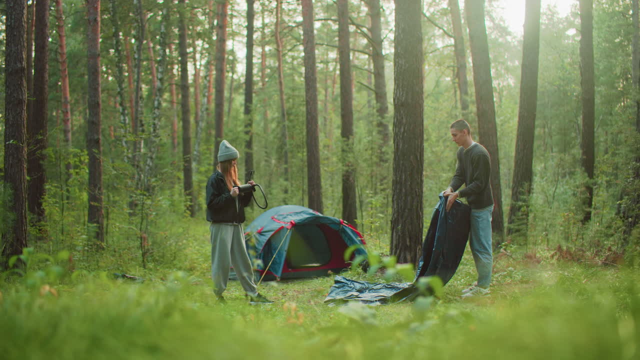 Young woman holding hose stands in forest clearing watching man spray tent fabric near set-up tent, both surrounded by tall trees and morning sunlight in calm outdoor camping environment