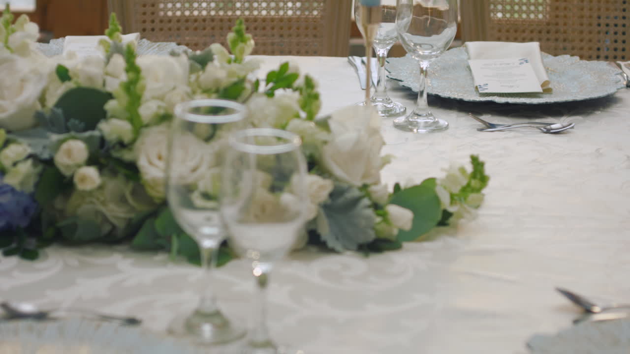 detailed clip of an elegantly decorated wedding table. It features stemware, a white tablecloth, plates, cutlery, cloth napkins, a delicate floral arrangement, and a personalized invitation.