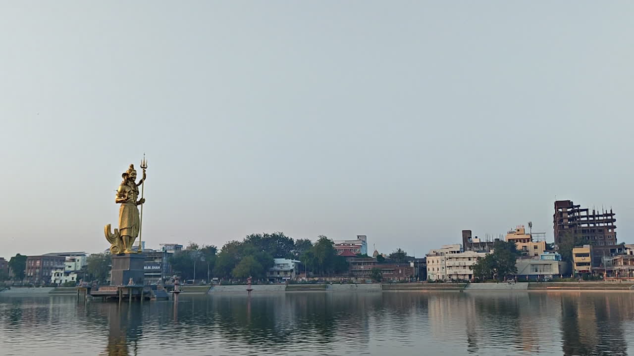 Cattura la bellezza di una vista laterale della magnifica statua dorata di Lord Shiva al lago Sursagar a Vadodara mentre cala il crepuscolo, proiettando un bagliore incantevole sulle acque serene.