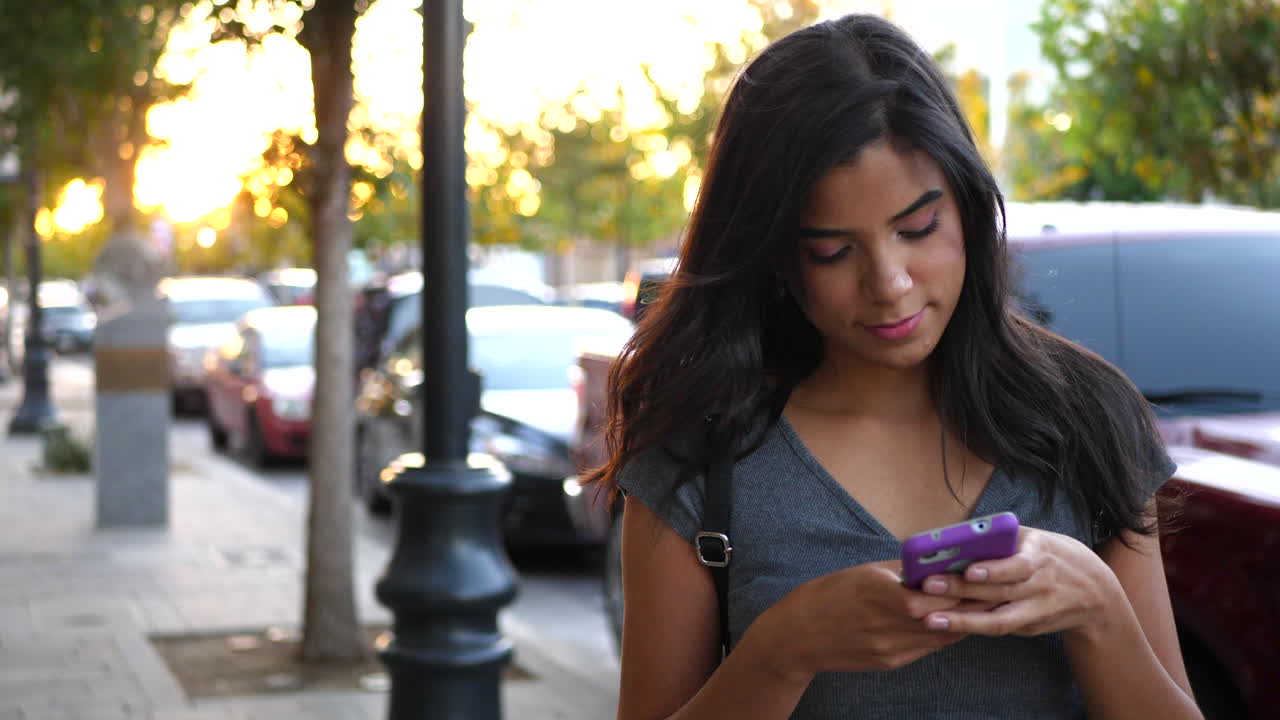 Una giovane donna cammina su un marciapiede cittadino, inviando messaggi o usando i social media sul suo smartphone al tramonto. SLOW MOTION.