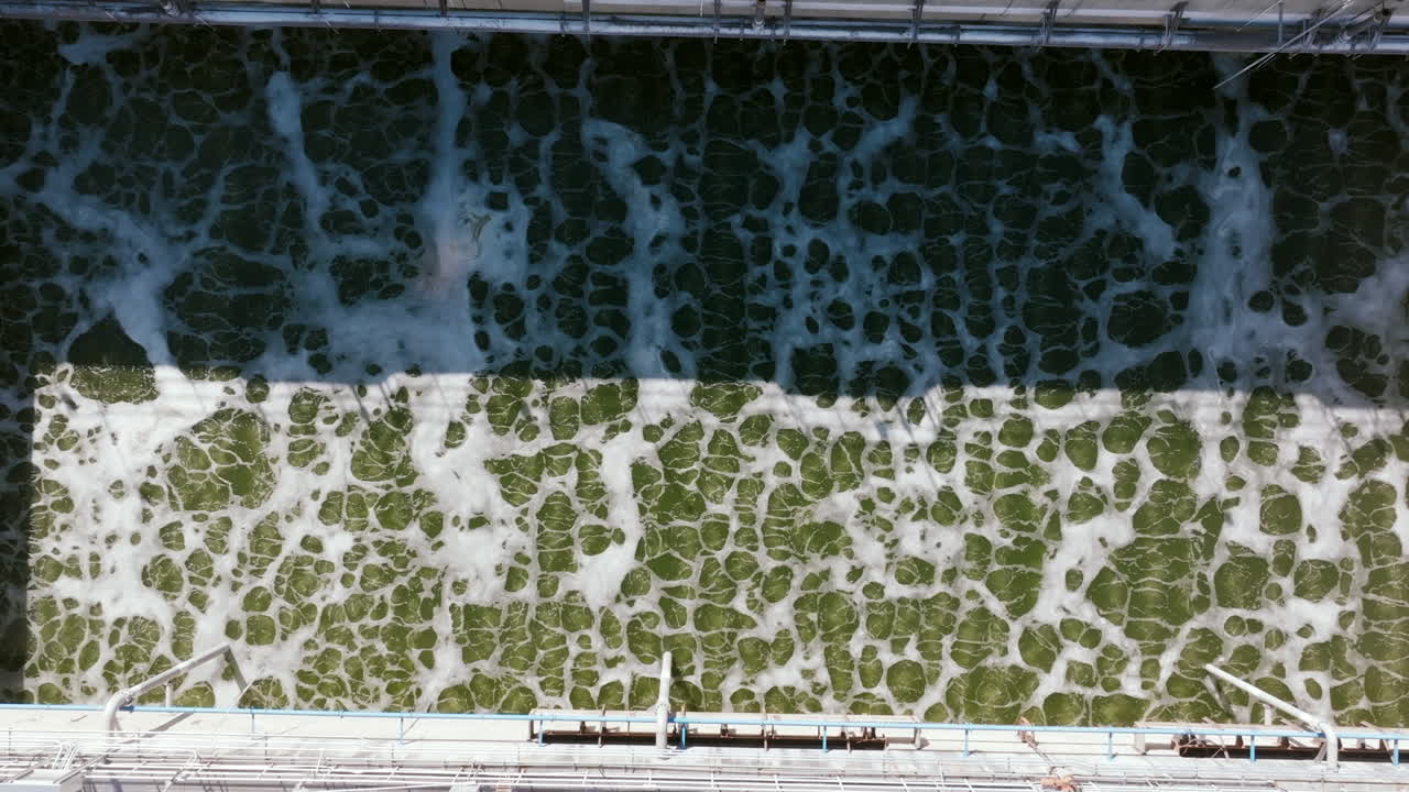 A top-down shot displays two separate treatment tanks—one filled with green, aerated water and the other with beige, foamy sediment—divided by a concrete walkway