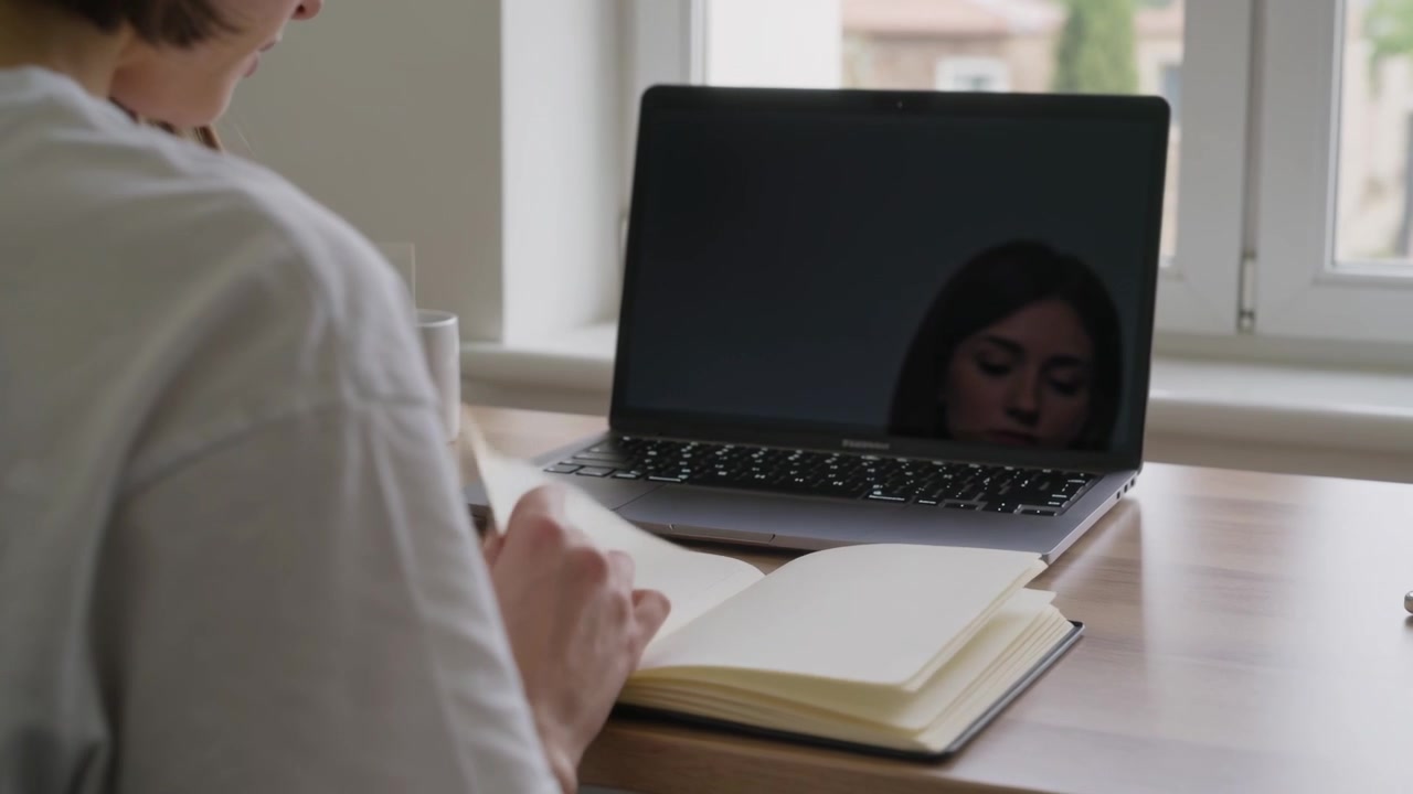 Flipping pages after noticing blank notebook, woman organizing notes at desk with laptop reflection