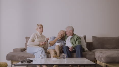 group of three senior people watching tv, talking and eating caramel popcorn while sitting on sofa at home