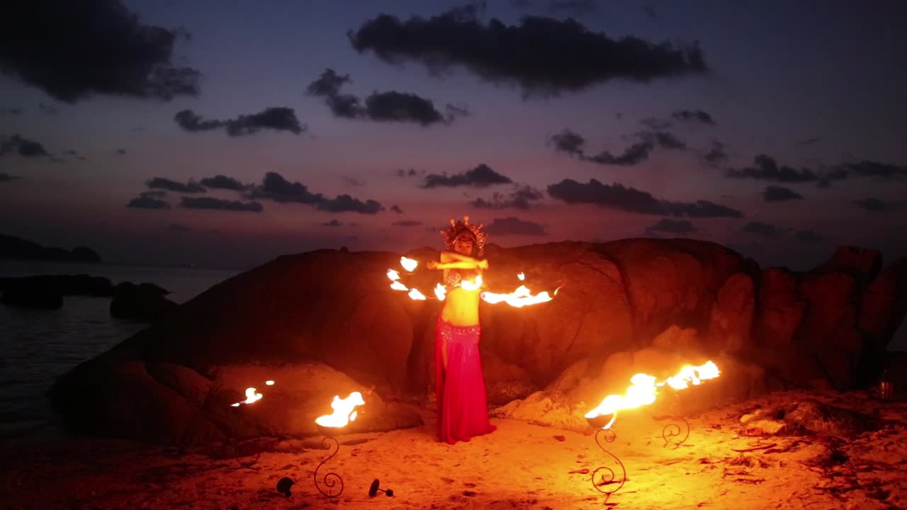 Erotic and sensual belly dancer in red dress, performing with lotus shaped fire props on the beach during golden hour.