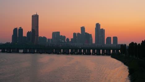 a beautiful sunset casts a warm orange glow over the yeouido skyline and hangang river in seoul, south korea, as a subway train crosses a railway bridge