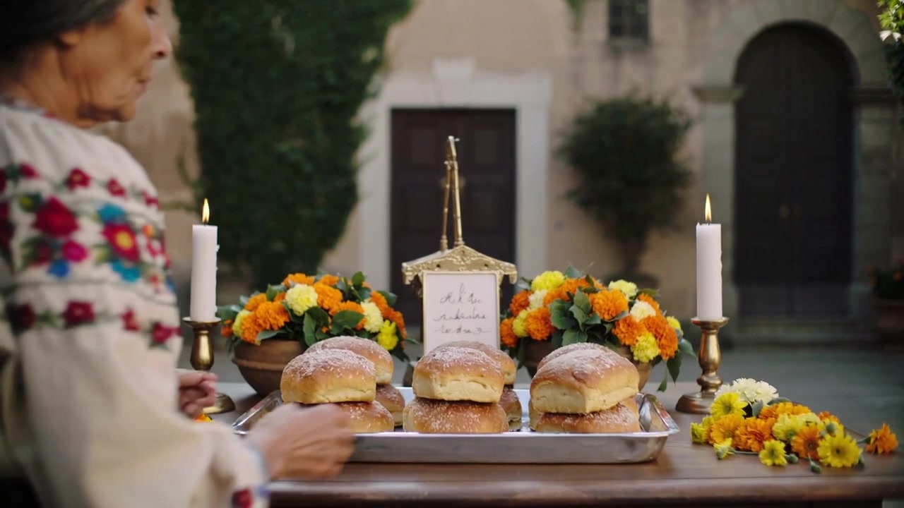 Arriving Hispanic woman wearing blouse placing breads flowers, brass candle on table making altar