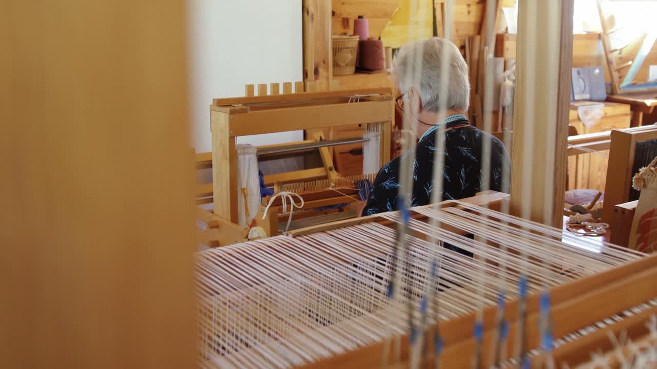 Rear view of old caucasian senior woman preparing and sitting at handloom machine in a workshop 4k