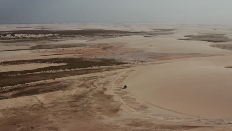 Aerial:-A-truck-with-kitesurfers-traveling-through-the-dunes-of-Lencois-Maranhenses-in-Brazil,-during-dry-season