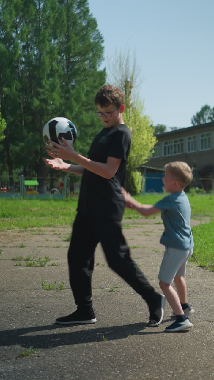 dos hermanos están jugando al fútbol juntos en una carretera pavimentada, con un edificio y un campo de hierba verde exuberante en el fondo