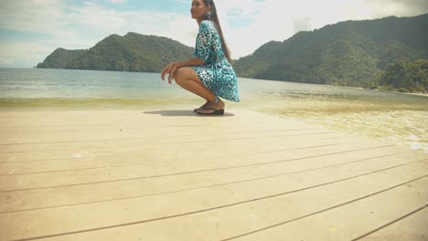 Una-Modelo-Posando-Sentada-En-Un-Embarcadero-Con-La-Playa-Y-Las-Montañas-Al-Fondo