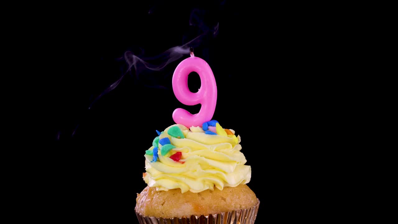 A single cupcake with yellow frosting and a pink number nine candle is blown out, with smoke rising, against a solid black background under studio lighting