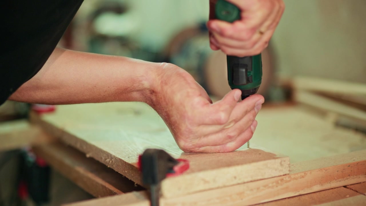 Close-up of a craftsman using a cordless screwdriver to fasten a screw into a wooden plank in a workshop. Authentic DIY woodworking footage for construction and assembly projects