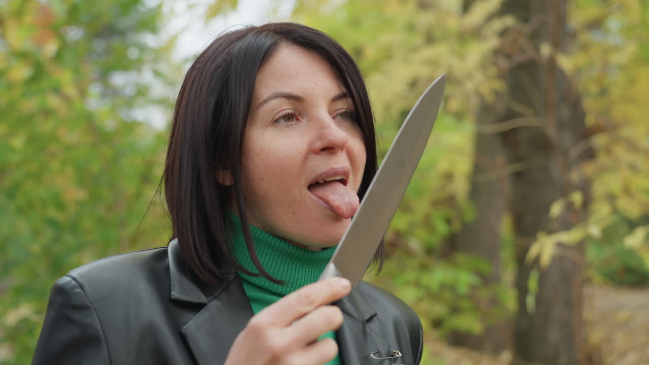 Donna con giacca di pelle che tiene in mano un coltello, dolcevita verde, foresta autunnale sullo sfondo. Gesto giocoso e rischioso della lingua vicino alla lama, sguardo rivolto verso l'alto, verso la chioma, primi piani che mostrano lentiggini e dettagli sottili.