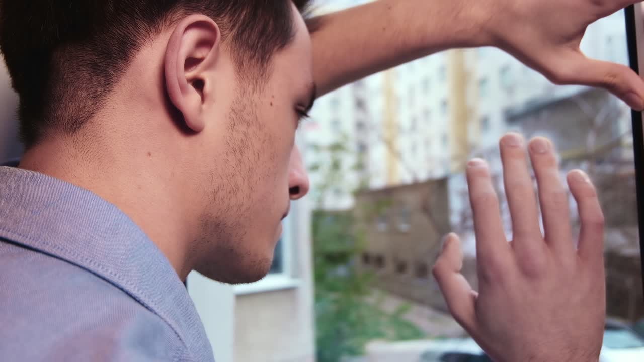 Bored man in blue shirt looking desperate out of the window and put the palm on it. Close up. Quarantine and Corona Virus idea