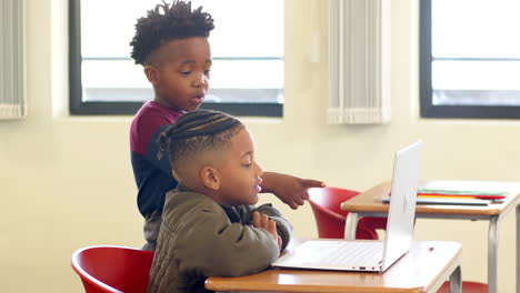 in school, young african american boy pointing at laptop, helping classmate with assignment