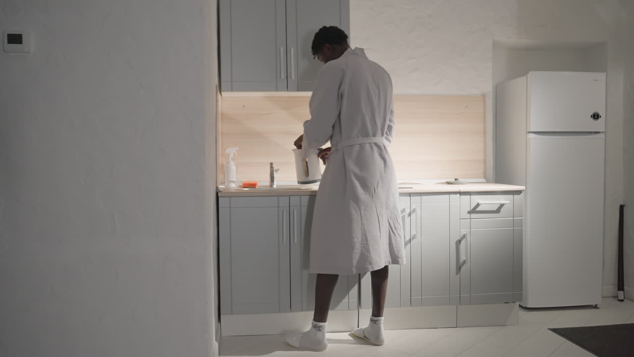 Guy in bathrobe standing in modern kitchen, pouring water into kettle. Bright, soft light fills the kitchen, creating a calm and focused morning atmosphere for preparing a hot drink
