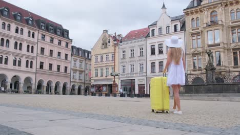 a slender french tourist in a beautiful white dress walks with a yellow suitcase in the center of the old town. tourist woman against the background of beautiful european architecture
