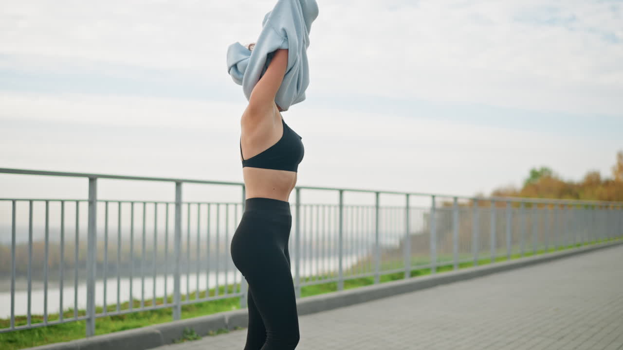 Lady standing in front of iron fence, removing her blue hoodie under sunny weather with a river background. Ideal for fitness, outdoor, and lifestyle moments in a bright, active environment