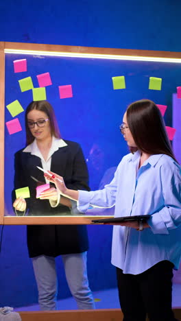 brunette women stand on both sides of the glass board. lady in black jacket takes the stickers off the board. brainstorming concept. vertical video.