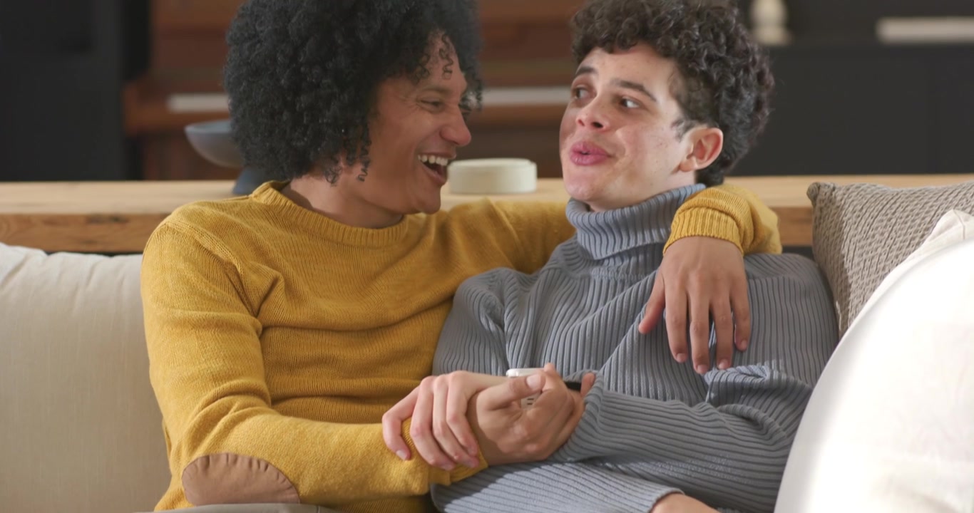 African American men laughing, holding hands while gesturing at offscreen TV on sofa with remote