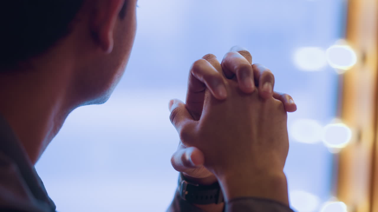 Rear view of young man sitting by window with clasped hands raised near face, softly lit by daylight, capturing moment of introspection, peaceful silence, and emotional stillness in thoughtful atmosphere