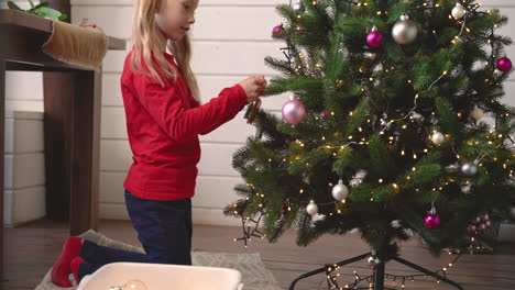 niña rubia de arrodillada en el suelo colgando decoración navideña en el árbol de navidad