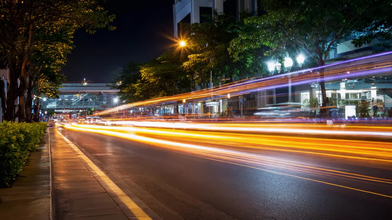 A Nighttime Urban Scene Captured in Motion, Featuring Vibrant Light Trails from Passing Vehicles Along a Quiet Road Surrounded by Lush Greenery and Illuminated Structures in a Bustling City Environment