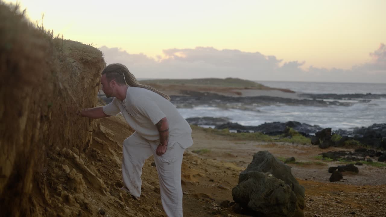 A man in white clothing carves into a dirt wall along a rocky coastal path, leaning closer as ocean waves crash behind him in the warm morning light