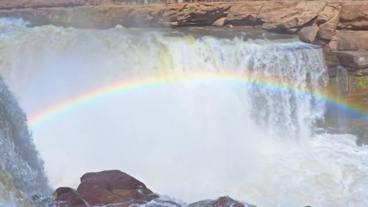 Panorâmica detalhada dos rápidos agitados e carregados de silte do Rio Amarelo até a névoa e o arco-íris na beira da Cachoeira Hukou. Apresenta a água marrom única e o leito rochoso do rio