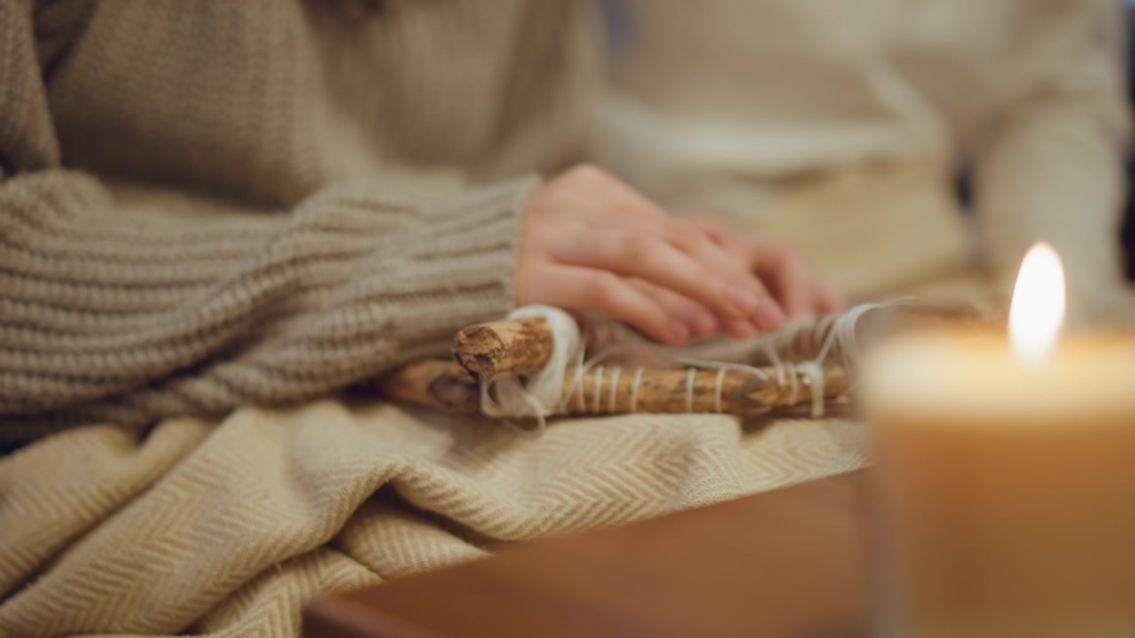 Close up of lit candle on wooden surface with soft warm light. Background shows person in cozy knitwear engaging gently with natural fiber craft creating calm indoor scene with neutral tones