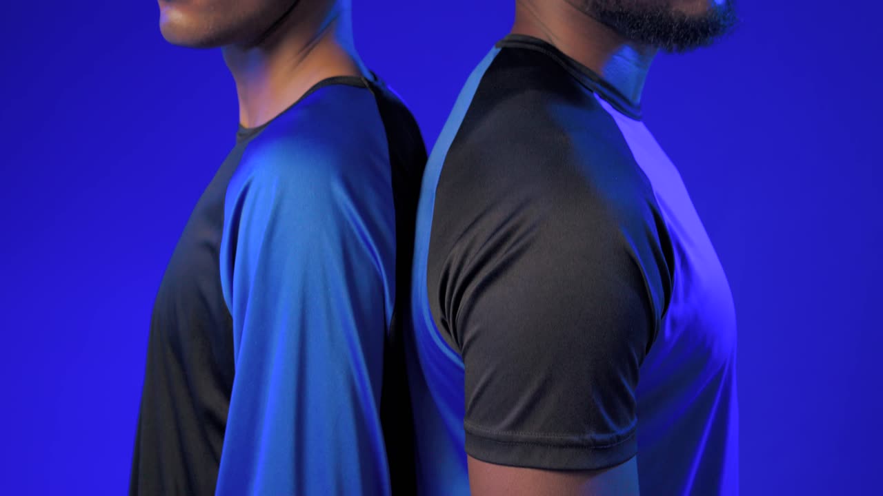 Dos hombres afrodescendientes con camisetas deportivas, espalda con espalda, con la cámara elevándose en un set azul para mostrar un guante de portero.