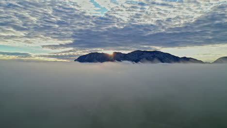 aerial view of sun rising behind a mountain, piercing through a blanket of clouds at dawn