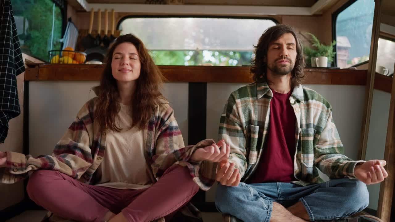 Portrait of a meditating couple, a brunette girl in a plaid shirt and pink pants along with her boyfriend in a Green plaid shirt and a red T-shirt sitting in the lotus position and meditating in their country house or trailer during their picnic outside the city in the summer