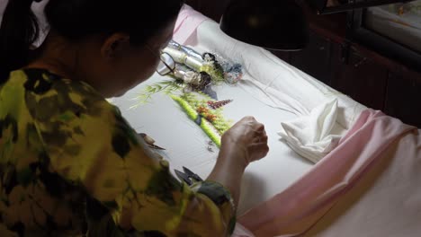close-up view of a woman skillfully embroidering a detailed landscape scene.